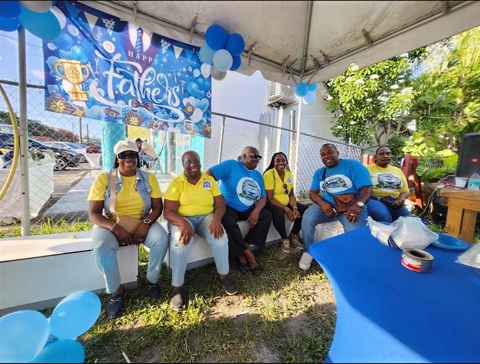 Volunteers in yellow and blue shirts sit on a bench under a Happy Father's Day banner and balloons