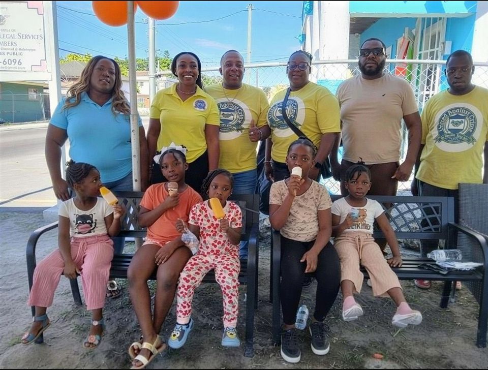 Volunteers in yellow and blue shirts stand behind children eating popsicles on benches at the Thanksgiving feeding